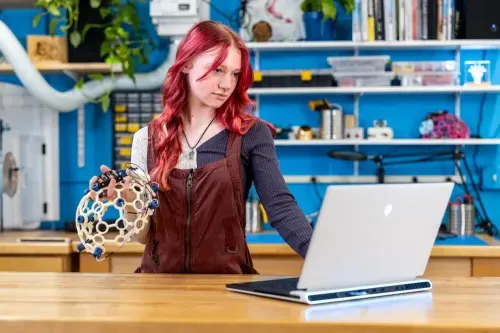 A U N E student holds up a brain wave helmet and works on their laptop